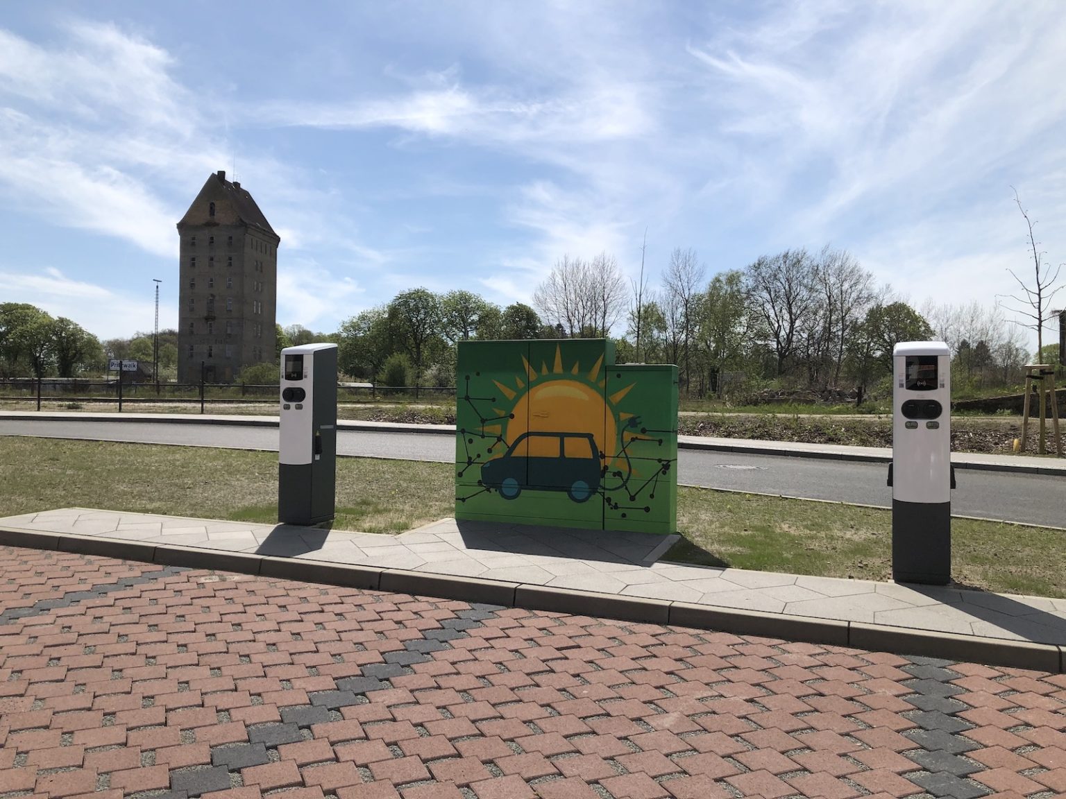 2 E-Ladesäulen am Bahnhofsvorplatz links und rechts von einem bemalten Stromkasten. Das Bild zeigt ein Auto vor einer Sonne.
