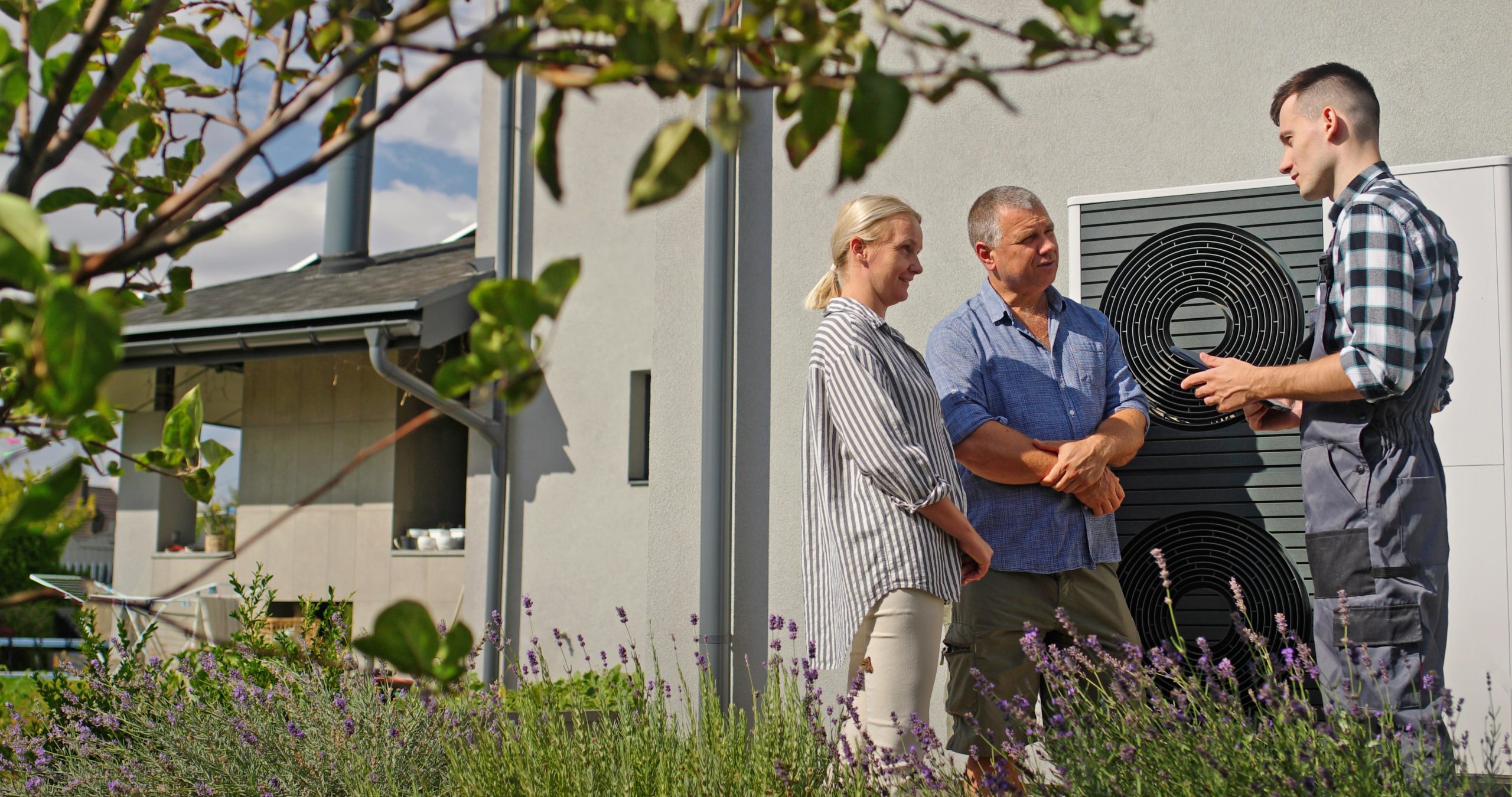 Ein Techniker spricht mit zwei Personen, während sie draußen neben einer Wärmepumpe stehen.